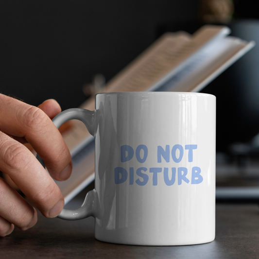 White mug with 'DO NOT DISTURB' text held by a hand on a desk.