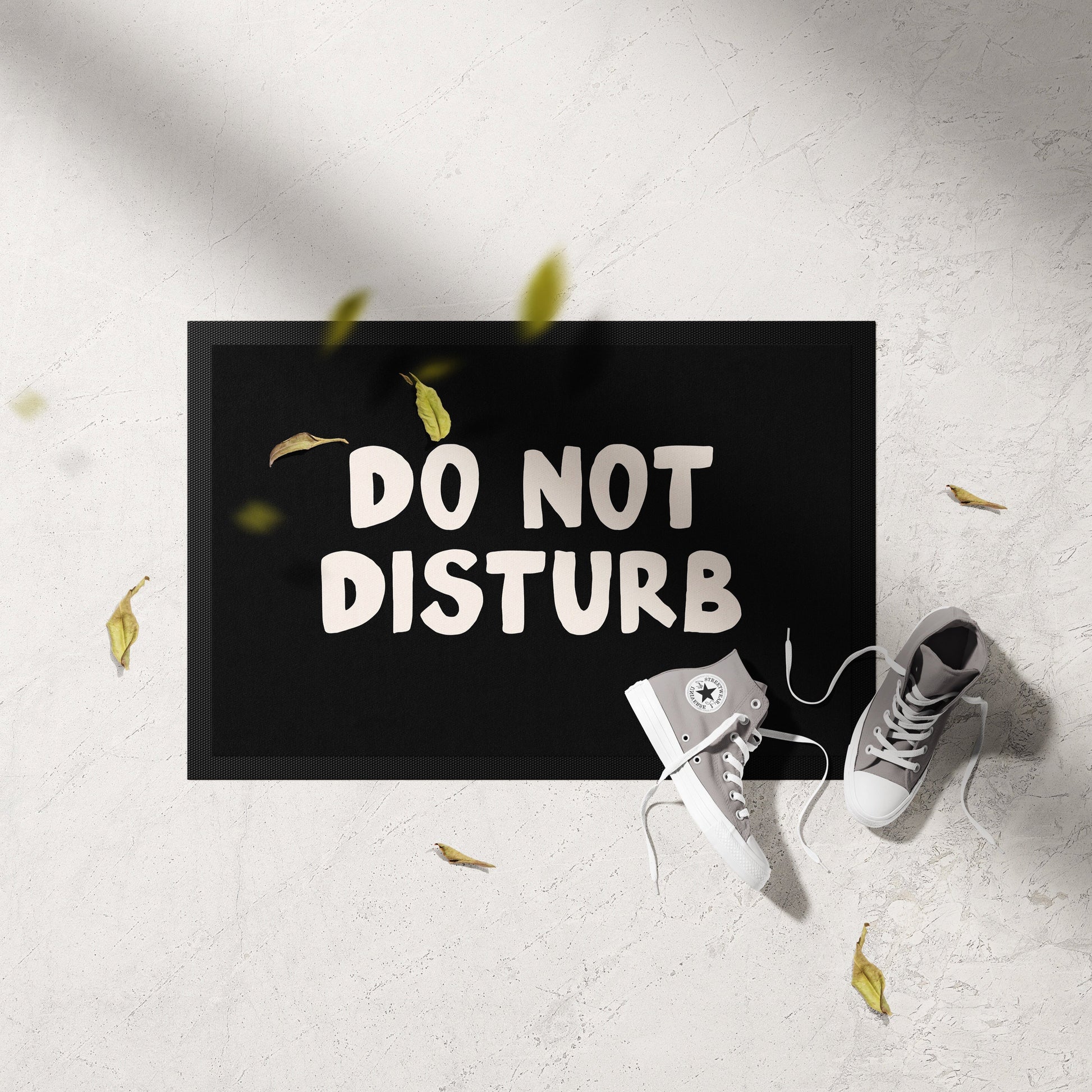 Doormat with 'DO NOT DISTURB' text on a light gray floor with white sneakers and yellow leaves.